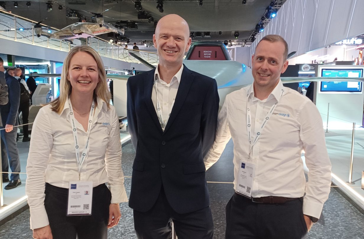 From left to right: Sarah, Mike and Phil stand smiling, dressed in smart clothing, in front of an exhibition stand