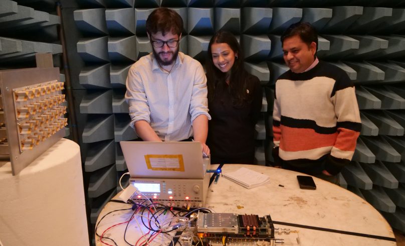 Sam, Rucha, Krishna (left to right) stand smiling in front of a laptop that is open and operating an electrical assembly radio unit on the table in front of them. They are in an anechoic chamber with a blue background